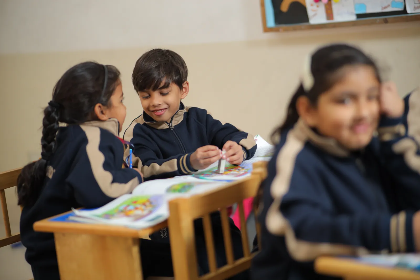 Two learners sharing a picture book at a wooden desk, heads together.
